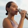 A woman drinks from a metal bottle on a sunny day by the beach, wearing a sports bra.